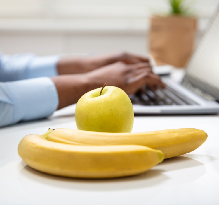 foto de uma banana e maça e no fundo uma pessoa digitando no laptop