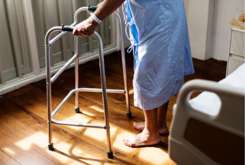 foto de uma idosa vestindo um avental em pé (com apoio de um andador), do lado da cama de um hospital